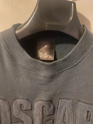 Mörkgrå sweatshirt Oscar Jacobson M - Snygg mörkgrå sweatshirt från Oscar Jacobson i storlek M. Tröjan har ett stort broderat OSCRAR-tryck på bröstet och klassisk rund hals. Mjukt material med ribbade muddar vid ärmslut och nederkant, perfekt för chill dagar. Sitter mer som S