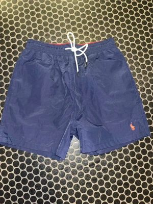 Blå badshorts Polo Ralph Lauren  - Snygga marinblå badshorts från Polo Ralph Lauren. Dessa shorts Passar mer som M/S i storlek. med vit snörning i midjan och klassisk röd broderad logga på benet. Shortsen har meshfoder, elastisk midja och praktisk bakficka. Perfekta för stranden eller poolen!