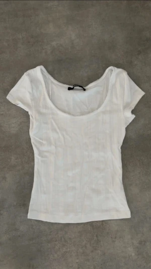 Vit ribbad topp med rund hals - Säljer en vit ribbad topp med rundad hals och korta ärmar. Toppen har en slim passform och diskreta vertikala ränder i materialet. Perfekt för dig som gillar enkel och clean stil. Superenkel att matcha med jeans eller kjol!