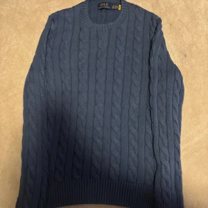 Blå kabelstickad tröja Polo Ralph Lauren - Snygg blå kabelstickad tröja från Polo Ralph Lauren. Tröjan har rund hals, ribbade muddar och ett klassiskt kabelmönster över hela plagget. Perfekt för dig som gillar stilrena och tidlösa plagg med premiumkänsla.