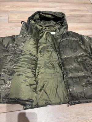 Gucci mörkgrön pufferjacka med huva - Säljer en fet mörkgrön pufferjacka från Gucci med GG-logga över hela jackan. Jackan har huva, dragkedja framtill och två sidofickor med dragkedja. Materialet är glansigt och känns lyxigt, perfekt för kalla dagar. Jackan är långärmad och har en loose passform. M passar även S 