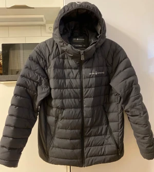 Sail racing  - Svart dunjacka från Sail Racing med huva och snygga quiltade paneler. Jackan har dragkedja framtill, två sidofickor med dragkedja och logga på bröstet. Materialet är nylon och dun, vilket gör den både lätt och varm. Perfekt för kalla dagar och stilren look.