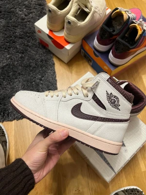 Nike Air Jordan 1 A Ma Maniére High - Nike Air Jordan 1 A Ma Maniére High i ljusgrå mocka med vinröda detaljer och mönstrad krage. Swoosh och häl i vinrött reptilmönster, beige snörning och platt sula i ljusrosa. Klassisk Air Jordan-logga på sidan och specialtag vid snörningen.