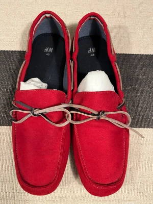 Röda mockaloafers från H&M strl 43 - Snygga röda loafers från H&M i storlek 43. Skorna är gjorda i mocka med dekorativt snöre runtom och rosett framtill. De har en klassisk seglarsko-look med platt sula och fyrkantig tå. Perfekta för dig som vill sticka ut med färg på fötterna. Använd bara 1 gång.