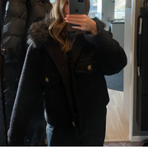 Svart med pälskrage - Säljer en svart jacka med fluffig pälsig krage och snygga bruna knappar framtill. Jackan har två stora fickor med lock och är croppad i modellen. Perfekt för dig som vill ha en cool och mysig look i höst och vinter.