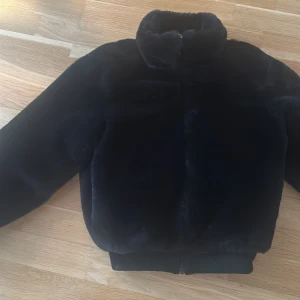 Svart pälsjacka med dragkedja - Säljer en svart pälsjacka med hög krage och croppad passform. Jackan har en snygg ringformad dragkedja framtill och är riktigt mjuk och fluffig. Perfekt för dig som vill ha en stilren och cozy look under kyliga dagar.