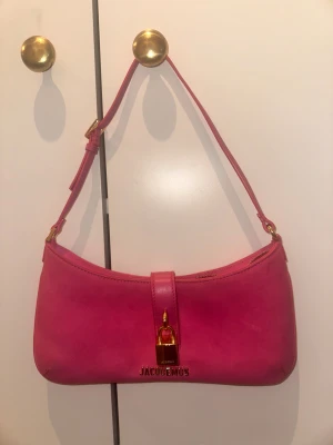 Rosa axelväska från Jacquemus - Säljer en snygg rosa axelväska från Jacquemus med guldfärgat lås och logga framtill. Väskan har en stilren, avlång form och justerbar axelrem. Materialet är mjukt skinn och detaljerna i guld ger en lyxig vibe. Perfekt för dig som vill sticka ut med en färgklick. Ny pris 9900kr.