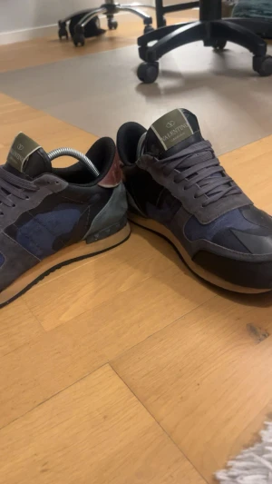 Valentino Garavani Rockrunner sneakers - Säljer ett par Valentino Garavani Rockrunner sneakers i mörkblått och grått med coolt kamouflagemönster. Skorna har snörning, platt sula och detaljer i mocka, skinn och textil. Ikonisk design med kontrastfärgad häl och box i camo-print med guldlogga.