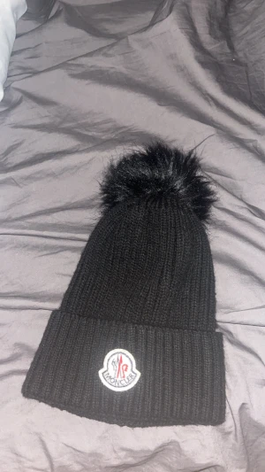 Svart Moncler mössa med pälsboll - Snygg svart mössa från Moncler med stor fluffig pälsboll på toppen och klassisk Moncler-logga framtill. Mössan är ribbstickad och har uppvikt kant för extra stilpoäng. Perfekt för dig som vill ha en trendig och varm accessoar i vinter.