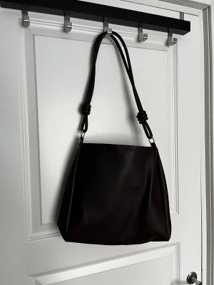 Brun axelväska med knutdetaljer - Stilren brun axelväska i skinnimitation med mjuk, fyrkantig form. Väskan har breda axelband med coola knutdetaljer och silverfärgade metalldelar. Perfekt storlek för att få plats med allt du behöver och passar till många olika outfits.