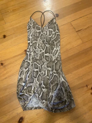 Snakeprint kortklänning från H&M - Supersnygg kortklänning från H&M Divided med ett coolt ormskinnsmönster i beige, grå och svart. Klänningen har tunna axelband och en figurnära passform som ger en trendig look. Perfekt för dig som vill sticka ut med ett djuriskt mönster.