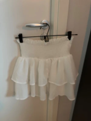 Vit volangkjol med smock - Supersöt vit kortkjol med bred smock i midjan och dubbla lager volanger. Kjolen är gjord i ett lätt och luftigt material som ger en flowy känsla. Perfekt för dig som gillar romantisk och feminin stil. Passar till crop tops och sneakers för en chill look.