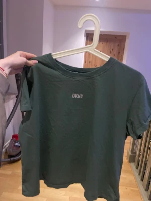 Grön t-shirt från DKNY - Snygg grön t-shirt från DKNY med klassisk rund hals och diskret logga framtill. Modellen är enkel och har korta ärmar, perfekt för en chill och stilren look. Materialet är mjuk bomull som känns skönt mot huden.
