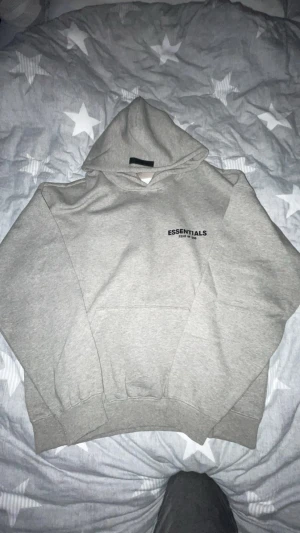 Grå Essentials Fear of God hoodie - Snygg grå hoodie från Essentials Fear of God med svart logotyp på bröstet och stor text på ryggen. Tröjan har huva, ribbade muddar och en klassisk känguruficka framtill. Perfekt för dig som gillar streetwear och vill ha en clean look.
