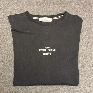 Svart Stone Island t-shirt med tryck - Svart t-shirt från Stone Island med ARCHIVIO-tryck på bröstet och ett stort guldigt tryck med text på ryggen. Rund hals och korta ärmar. Materialet är mjuk bomull och passformen är normal. Perfekt för dig som gillar streetwear och snygga detaljer. Nypris 2500