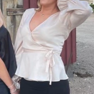 Champagnefärgad omlottblus med knyt - Säljer en champagnefärgad omlottblus med v-ringning och långa ärmar. Blusen har ett fint knyt i midjan och lätt glansigt material som ger en lyxig känsla. Perfekt för dig som gillar eleganta och trendiga toppar med feminin touch.