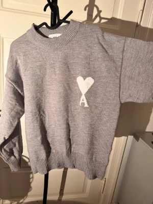 Grå stickad tröja från Ami - Snygg grå stickad tröja från Ami med ribbad krage, muddar och nederkant. finns ett vitt hjärta med bokstaven A. Tröjan har en loose passform och långa ärmar, perfekt för dig som gillar stilrena detaljer och vill ha något som sticker ut.