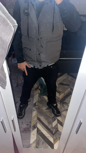 Grå dunväst från Canada goose  - Snygg grå dunväst från Canada goose med svarta detaljer och emblem på bröstet. Västen har stora fickor framtill, knappar och dragsko vid kragen. Perfekt att slänga över en hoodie eller tröja för en chill streetstyle-look.