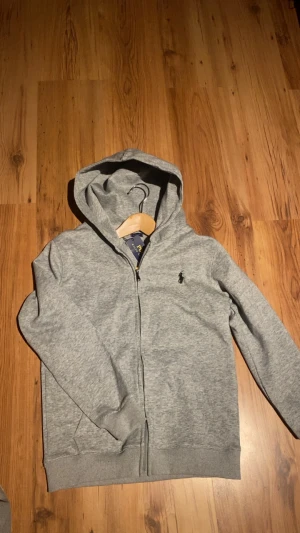 Grå hoodie från Polo Ralph Lauren - Snygg grå hoodie från Polo Ralph Lauren med dragkedja och huva. Klassisk svart logga broderad på bröstet. Mjuk bomullsmix, ribbade muddar och fickor framtill. Perfekt för chill dagar och streetwear-stil.