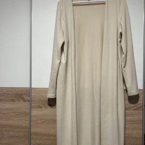 Beige lång cardigan med lång ärm - Säljer en stilren beige lång cardigan med lång ärm. Cardiganen har en enkel och rak design utan synliga detaljer eller mönster, vilket gör den superenkel att matcha med olika accessoarer. Materialet känns mjukt och bekvämt mot huden.