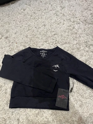 Svart långärmad träningströja Peach XS - Svart långärmad träningströja från Peach i storlek XS. Tröjan har en stilren design med diskret logga på bröstet och är tillverkad i ett stretchigt material av 90% nylon och 10% elastan. Perfekt för gymmet eller andra sportaktiviteter.