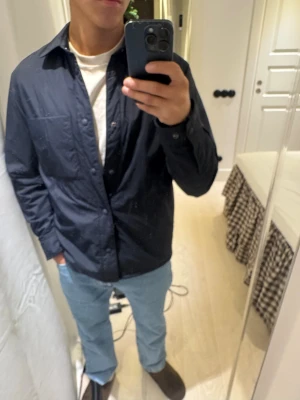 Mörkblå quiltad overshirt från ARKET - Quiltad overshirt från ARKET i mörkblå färg, med tryckknappar framtill och en stor bröstficka. Jackan har en clean look med klassisk krage och långärmad passform. Materialet är syntet och ger en lätt och smidig känsla. Perfekt för lager-på-lager under höst och vår. Inte använd alls nypris 1300 mitt  pris 900