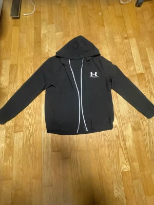 Svart vindjacka från D Project S - Svart vindjacka med huva från D Project i storlek S. Jackan har en vit dragkedja framtill och Under Armour-logga på bröstet. Långärmad och tillverkad i ett lätt syntetmaterial, perfekt för blåsiga dagar. Enkel och stilren design som funkar till streetwear.