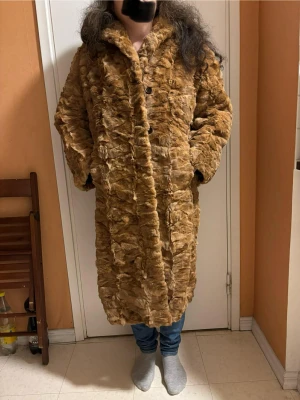 Brun lång äkta pälskappa - Säljer en lång och oversized pälskappa i brun ton med fluffig struktur och stora knappar framtill. Jackan har ett brunt, glansigt foder med snyggt bladmönster och två sidofickor. Perfekt för dig som vill sticka ut och hålla dig varm under kalla dagar. Den är vintage och det står varken storlek eller tillverkare. Jag är 162 cm och väger 63 kg. Kappan väger 3 kg.