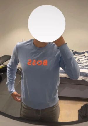 Ljusblå BOSS sweatshirt med orange tryck - Snygg ljusblå sweatshirt från BOSS med orange BOSS-tryck på bröstet. Tröjan har rund hals, ribbade muddar och är långärmad. Tillverkad i mjuk bomull som är skön mot huden. Perfekt för dig som gillar stilrena och cleana plagg med en färgklick.