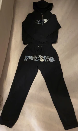 Svart Tracksuit från Trapstar - Svart tracksuit från Trapstar med fet logga på bröstet. Hoodien har huva, dragkedja och långa ärmar. Materialet är mjuk bomull, perfekt för chill dagar. Snygg streetwear-vibe och perfekt för de som gillar bold prints. Byxorna passar också S. Funderingar? Hör av dig 🤙