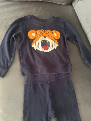 Mörkblå tröja med tigertryck Softer Days - Mörkblå långärmad tröja från Softer Days med stort tigertryck i orange och vitt framtill. Tröjan är i mjuk bomull och har ribbade muddar vid ärmslut och nederkant. Perfekt för dig som gillar coola djurmotiv och bekväma kläder.