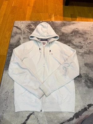 Tommy Hilfiger Denim - zip hoodie - Vit hoodie från Tommy Hilfiger Denim med dragkedja och huva. Tröjan har snörning med röda och marinblå detaljer samt broderad logga på bröstet. Mjuk insida och ribbade muddar. Perfekt för en chill och stilren look.