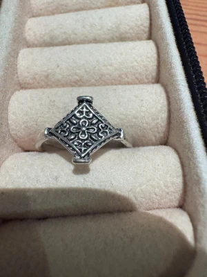 Silvrig ring med blommönster - Unik silvrig ring med fyrkantig topp och detaljerat blommönster. Ringen har en vintage vibe med snygga ornament och passar perfekt för dig som gillar boho eller retrostil. Materialet är metall med antik finish och mönstret ger extra karaktär.