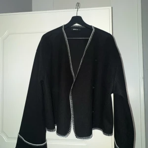 Svart kofta från Gina Tricot med vit kant - Svart kofta från Gina Tricot med kontrasterande vit kant längs öppningen och ärmsluten. Koftan har en loose passform, långärmade ärmar och är tillverkad i ett mjukt stickat material. Perfekt att slänga över en t-shirt för en chill look.