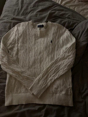 Vit kabelstickad tröja Polo Ralph Lauren - Säljer en vit kabelstickad tröja från Polo Ralph Lauren i storlek L. Tröjan har rund hals, långärmade ribbade muddar och det klassiska broderade Polo-logot på bröstet. Perfekt för dig som gillar clean och tidlös stil.