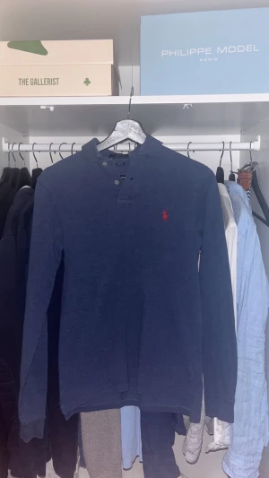 Polo Ralph lauren pikétröja  - Snygg marinblå långärmad pikétröja från Polo Ralph Lauren i slim fit. Tröjan har klassisk krage, knappar vid halsen och den ikoniska röda logon broderad på bröstet. Perfekt för dig som gillar stilrena och tidlösa plagg.