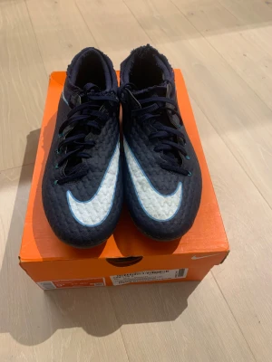 Nike Hypervenom phelon III - Nike Hypervenom fotbollsskor i mörkblå färg med stora vita Swoosh-loggor på ovansidan och sidan. Skorna har snörning och platt sula med blå dobbar för bra grepp på planen. Ovandelen är tillverkad i syntetmaterial med textur för bättre bollkontroll. Använda två gånger.