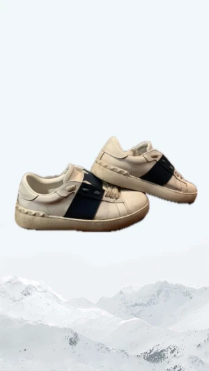 Valentino Garavani Open sneakers vit/blå - Snygga Valentino Garavani Open sneakers i vitt skinn med bred marinblå läderdetalj över snörningen. Klassisk rund tå, platt sula och ikoniska nitar längs hälen. Perfekta för dig som vill ha en stilren men ändå lyxig look.