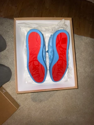 Blå Christian Louboutin sneakers - Självklart äkta allt på bilderna kommer med. Säljer ett par riktigt snygga blå sneakers från Christian Louboutin i mocka med ikoniska röda sulor och diskret logga på sidan. Skorna har snörning och en clean, modern look som sticker ut. Perfekta för dig som vill ha något unikt och exklusivt i garderoben.