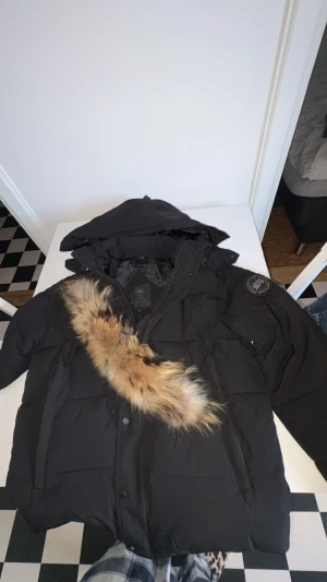 Svart dunjacka från Canada Goose - Säljer en svart dunjacka från Canada Goose med huva och avtagbar pälskant. Jackan har dragkedja, knappar och flera fickor. Perfekt för kalla vinterdagar och har en klassisk, puffig look med snygga detaljer. Jackan är storlek M/L