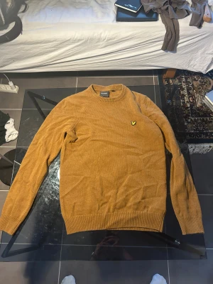 Brun stickad tröja från Lyle & Scott - Snygg brun stickad tröja från Lyle & Scott med klassisk rund hals och ribbade muddar. Tröjan har lång ärm och den ikoniska gula fågeln broderad på bröstet. Perfekt för dig som gillar stilrena och tidlösa plagg med en modern touch.