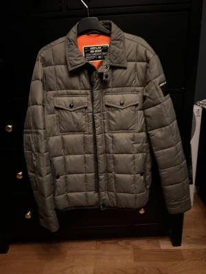 Grå quiltad pufferjacka från Replay - Säljer en grå pufferjacka från Replay med quiltad design och vattenavvisande yta. Jackan har två bröstfickor med knappar, Replay-logga på ärmen och orange foder. Perfekt för dig som vill ha en snygg och funktionell jacka med cool streetkänsla.