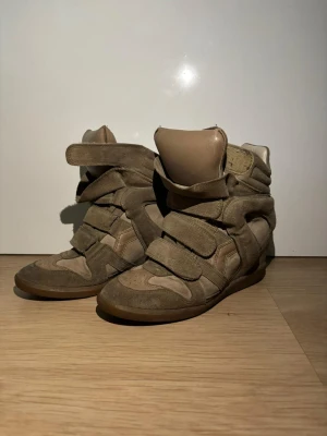 Beige Isabel Marant wedge sneakers - Coola wedge sneakers från Isabel Marant i beige mocka och skinn. Skorna har flera kardborreband, rund tå och en dold kilklack som ger extra höjd. Perfekt för dig som vill ha en sportig men ändå trendig look. Snygga detaljer och bekväm passform.