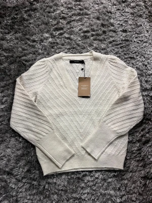 Vit stickad v-ringad tröja Vero Moda - Superfin vit stickad tröja från Vero Moda med v-ringning och diskret strukturmönster. Tröjan har långa ärmar, breda muddar och en snygg passform som känns både chill och stilren. Perfekt att matcha med jeans eller kjol.