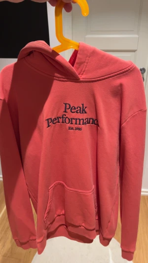 Korallrosa hoodie Peak Performance - Snygg korallrosa hoodie från Peak Performance med broderad logga på bröstet. Tröjan har huva, magficka och långa ärmar. Perfekt för chill dagar och har en skön, mjuk känsla tack vare bomullsmaterialet.
