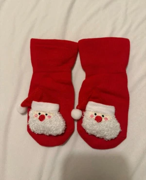 Röda julstrumpor med tomte - Supergulliga röda strumpor med fluffigt tomteansikte framtill och vita halkskydd under foten. Perfekta till julmyset och har en mysig design med broderade detaljer och en liten boll på tomteluva. Materialet känns mjukt och stretchigt, troligen bomull med syntet. 6-12 månader helt nya.