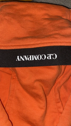 Orange hoodie från C.P. Company - Säljer en orange hoodie från C.P. Company i storlek M passar mer S. Tröjan har huva med snörning, klassisk linsdetalj på ärmen och en stor magficka. Materialet är mjuk bomull och passformen är normal. Perfekt för dig som gillar streetwear och vill sticka ut.