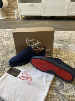 Mörkblå sneakers från Christian Louboutin - Snygga mörkblå sneakers från Christian Louboutin i mocka med klassisk röd sula och broderad logga på hälen. Skorna har rund tå, platt sula och snörning. Kommer med originalkartong och dustbag. Perfekta för dig som vill sticka ut med exklusiv stil. Lite använd men kan snygga till dem.