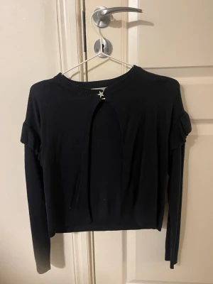 Svart kofta med stjärndetalj Gina Tricot - Svart kofta från Gina Tricot med lång ärm och snygga volangdetaljer på axlarna. Framtill finns en glittrig stjärnformad knapp som ger extra edge. Koftan är stickad och har en rund halsringning med ribbad kant. Perfekt för dig som gillar stilrena plagg med en twist.