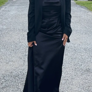 Svart lång satinklänning offshoulder  - Elegant svart långklänning i glansigt satintyg med offshoulder-ärm och draperad överdel. Klänningen har en figurnära passform upptill och faller snyggt längs kroppen. Perfekt för dig som vill sticka ut med en stilren och lyxig look. Har slit, sitter perfekt. (Kavajen ingår ej)